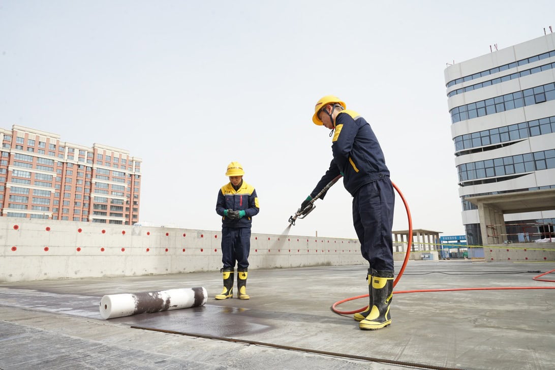 Two construction workers in yellow hard hats and blue uniforms on a rooftop, with one using a hose spray wand.