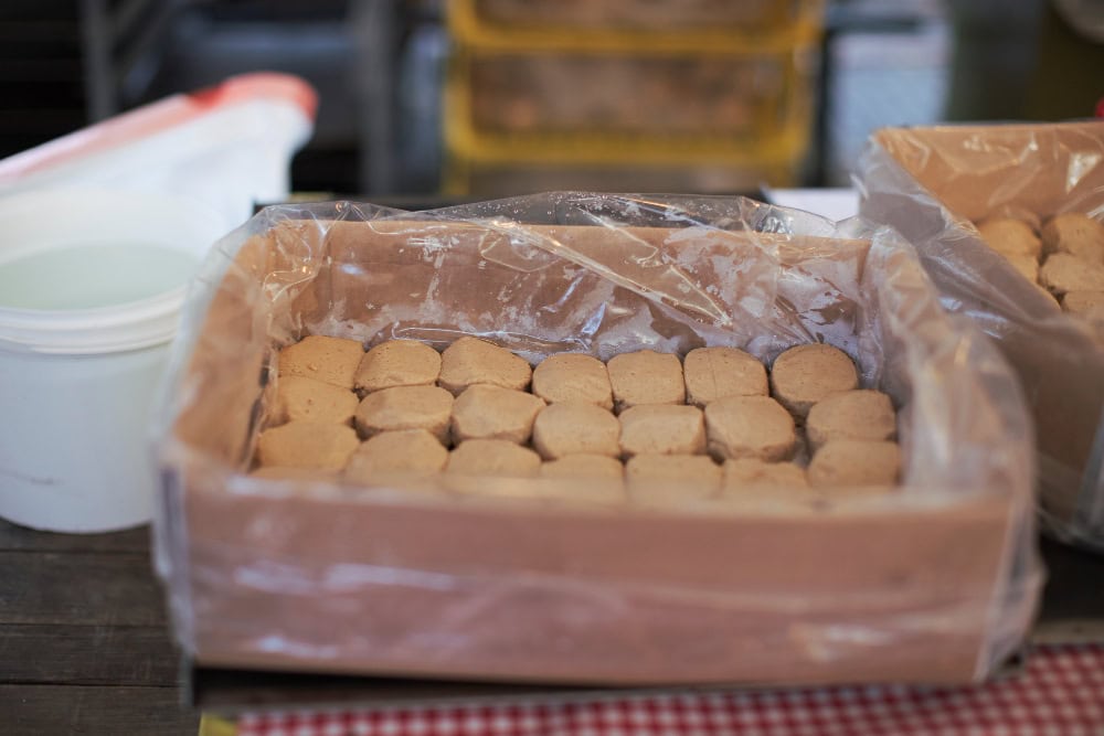 Tray of small beige cookies lined in rows inside a wooden box covered with plastic wrap.