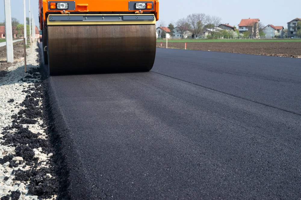 Road roller compacting fresh asphalt on a newly paved road with houses in the distance under a clear sky