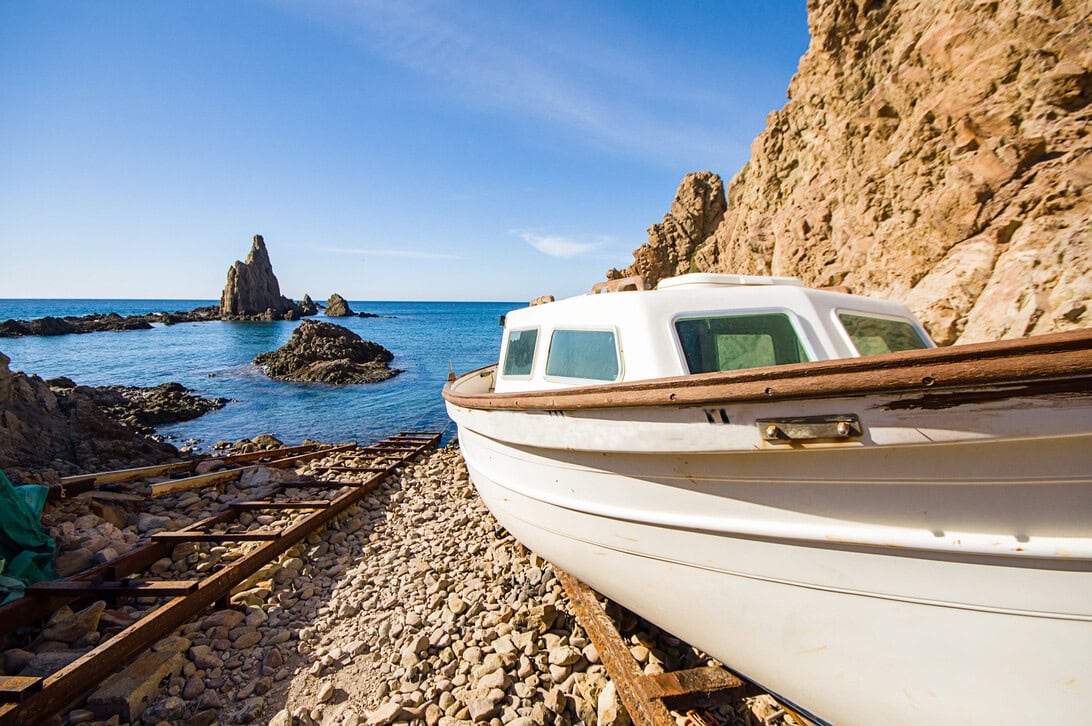 White boat on a rocky beach beside rusted rails with the ocean and cliffs in the background