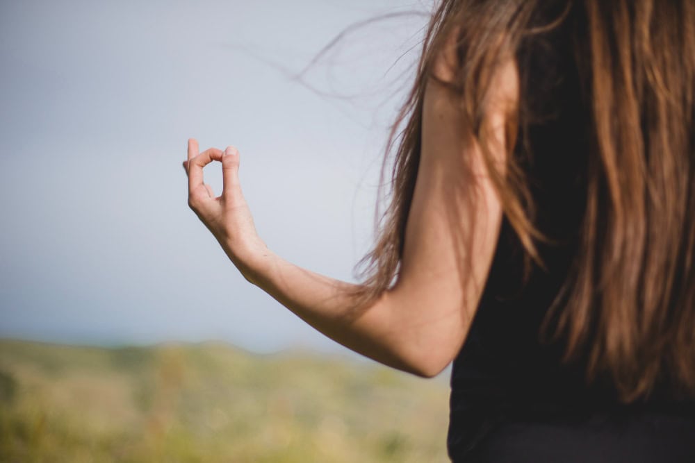 Back view of a person with long brown hair, wearing a black top, making an OK sign with their right hand outdoors.