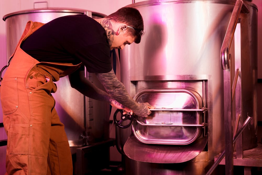 Man in tan overalls opens a large stainless-steel industrial kettle with a hand wheel on a metal door