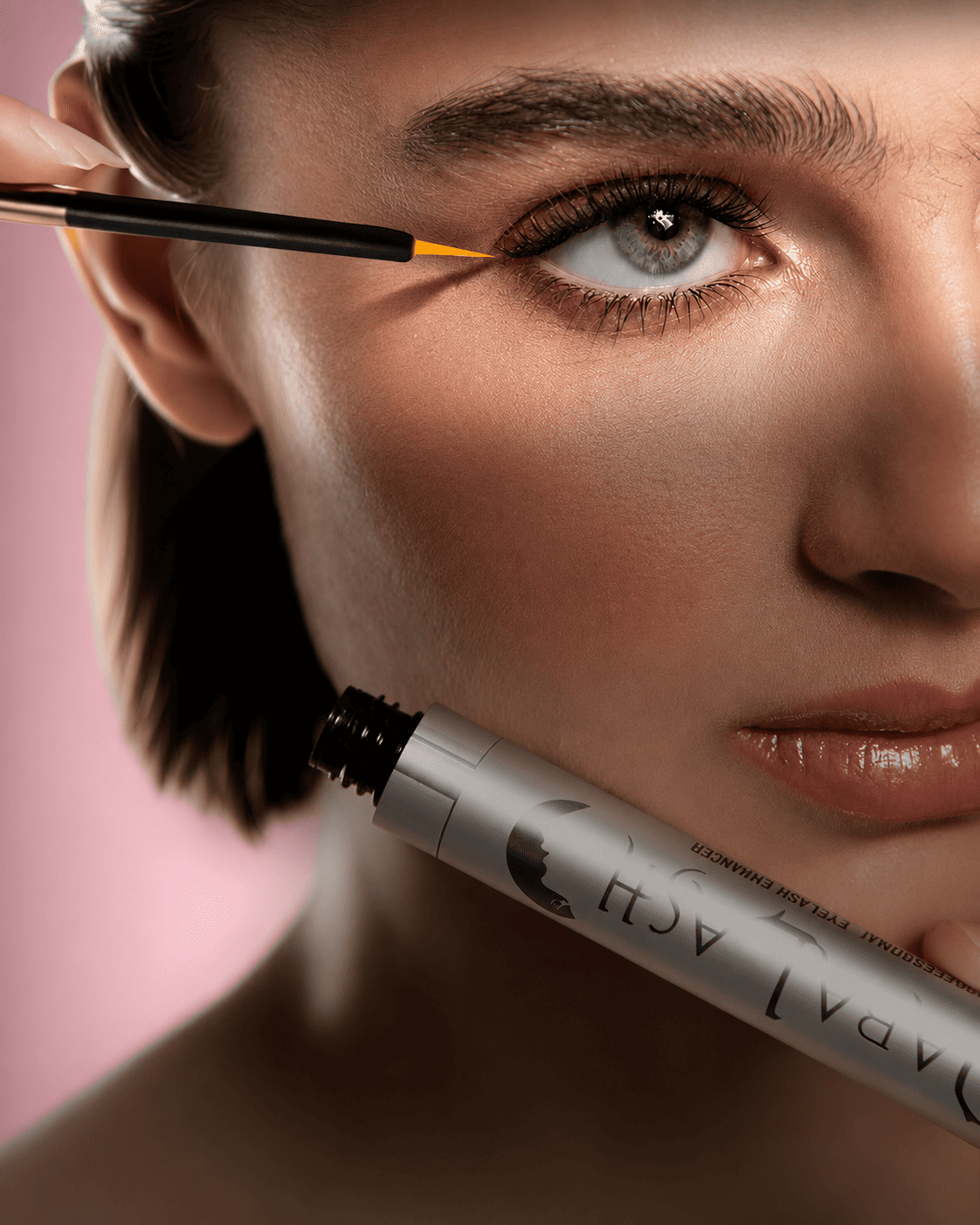 Close-up of a woman applying orange eyeliner along her upper lash line with a brush; a mascara tube rests near her chin.