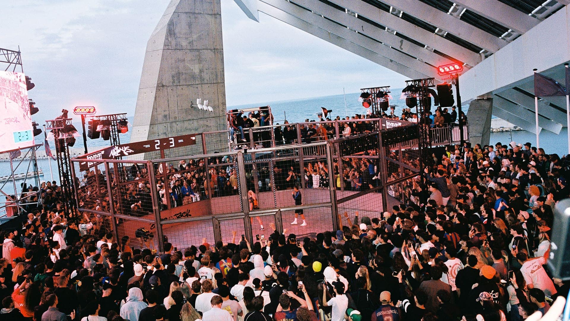 Crowd packed around a fenced performance area at a waterfront venue, with stage lights and the ocean in the background.