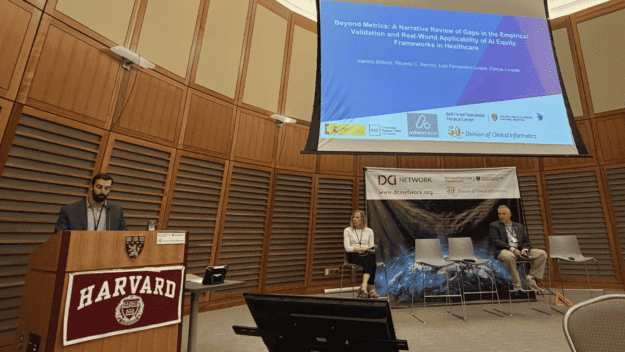 Speaker at a Harvard podium delivering a talk, with a large projection screen overhead and two panelists seated on stage to the right.