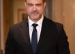Professional portrait: man in a black suit and navy tie standing in a hallway, looking at the camera.