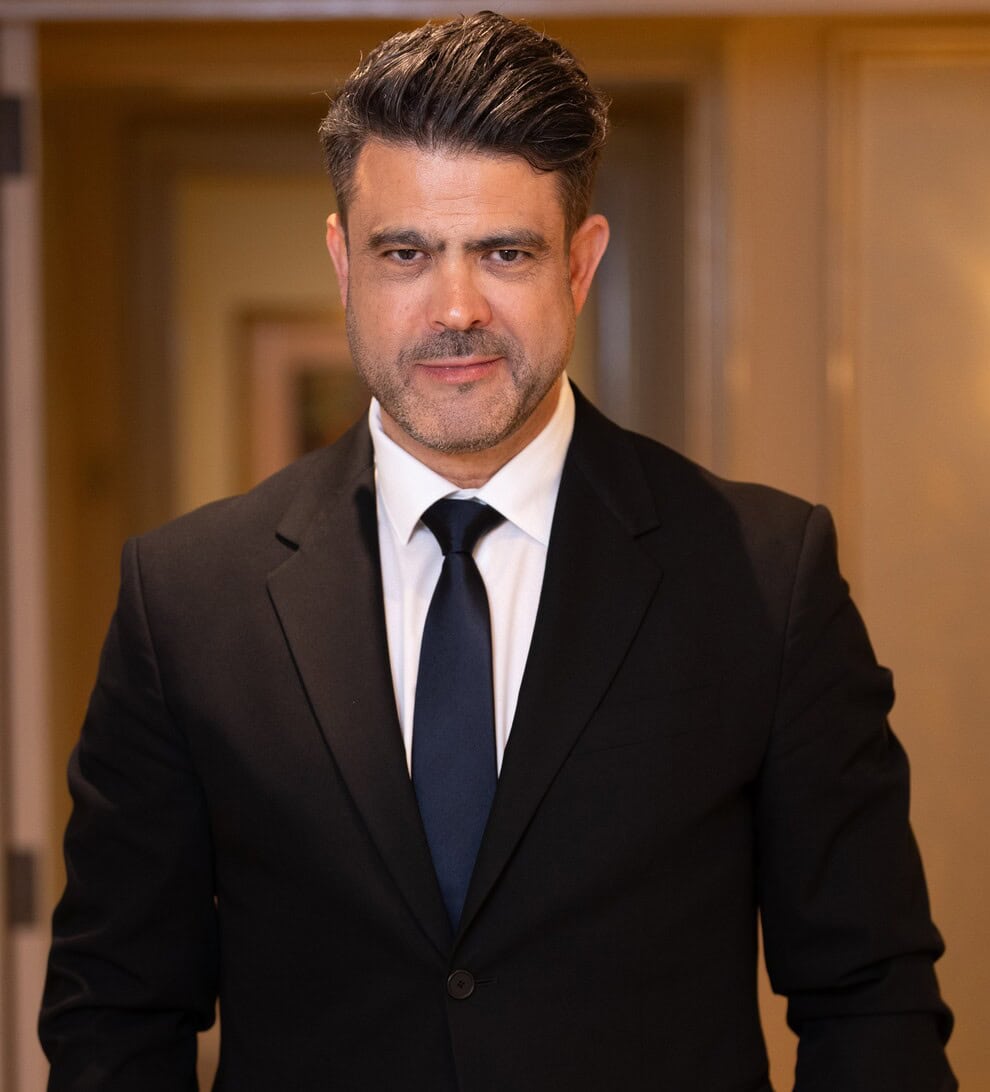 Professional portrait: man in a black suit and navy tie standing in a hallway, looking at the camera.