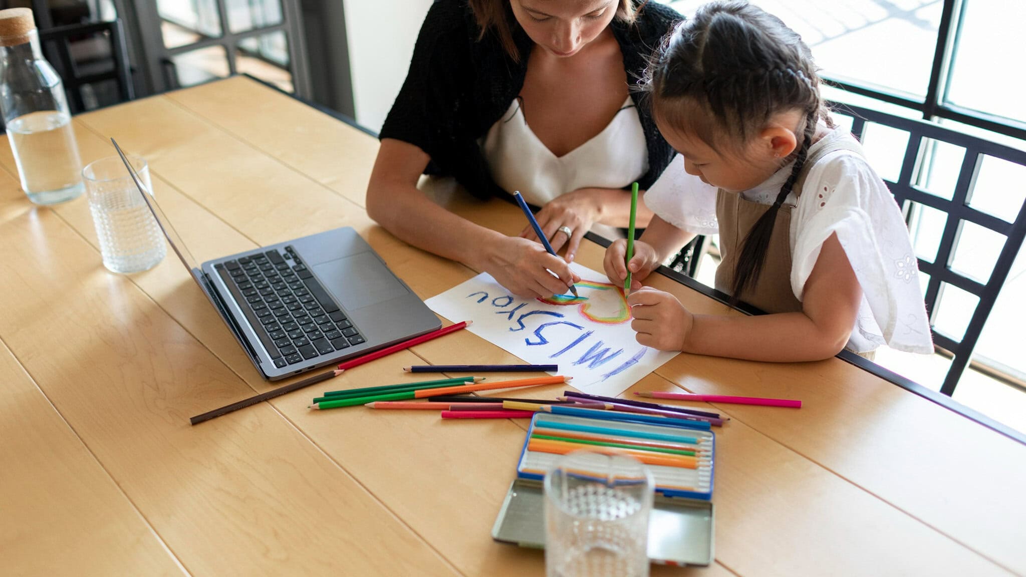 Woman and young girl drawing with colored pencils at a wooden table, laptop open nearby.