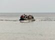 A group of people wearing life jackets crowded onto a black inflatable boat in the open sea, waves brushing the hull.