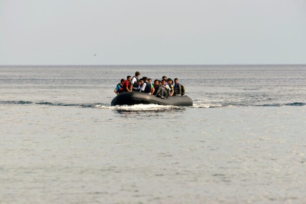 A group of people wearing life jackets crowded onto a black inflatable boat in the open sea, waves brushing the hull.