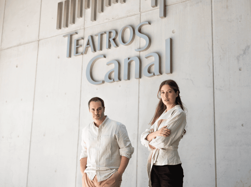 Two casually dressed adults, a man with hands in pockets and a woman with arms crossed, stand in front of a wall reading 'Teatros Canal'.