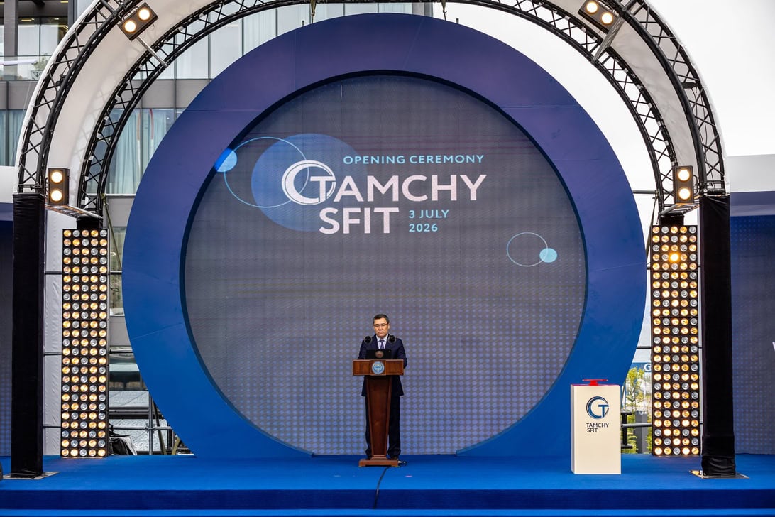 Man in a suit giving a speech at a wooden podium on a blue circular stage during an opening ceremony, with a large circular screen behind him that reads 'Opening Ceremony' and 'Gamchy Sift' (3 July 2026).