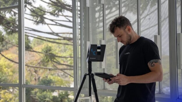 Man in a black shirt operates a tripod-mounted Matterport scanner, holding a tablet indoors by large windows.