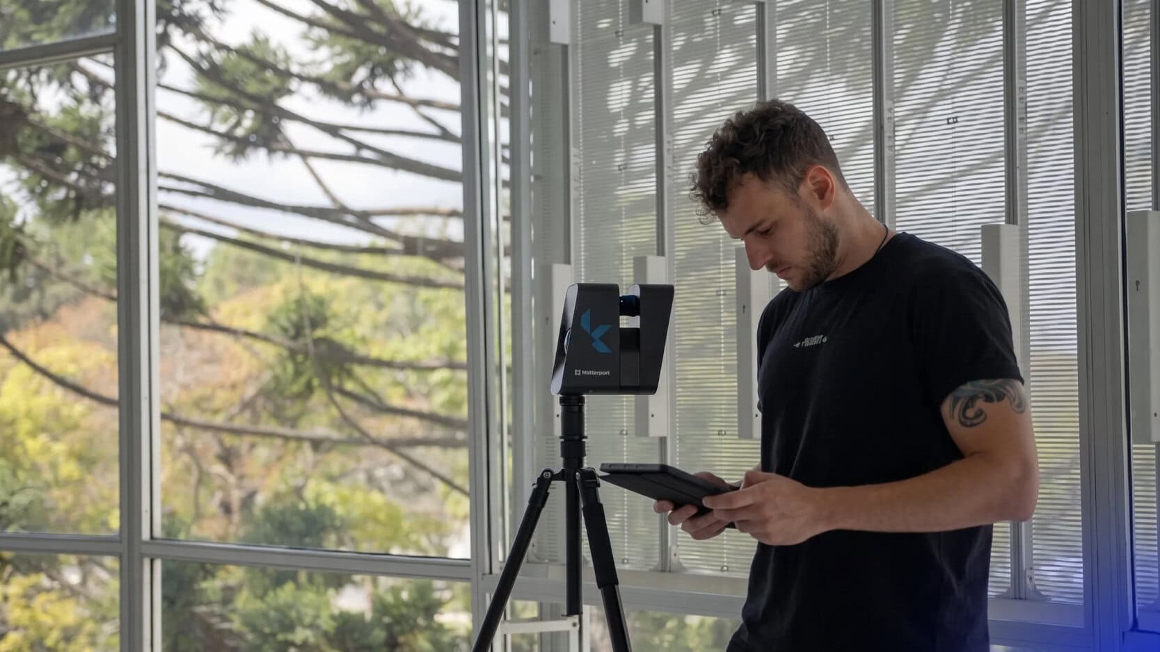 Man in a black shirt operates a tripod-mounted Matterport scanner, holding a tablet indoors by large windows.