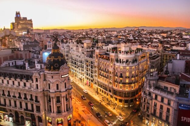 Sunset over a European city with ornate buildings and a prominent domed tower at a busy street corner, likely the Gran Via in Madrid, bathed in warm orange and pink hues.