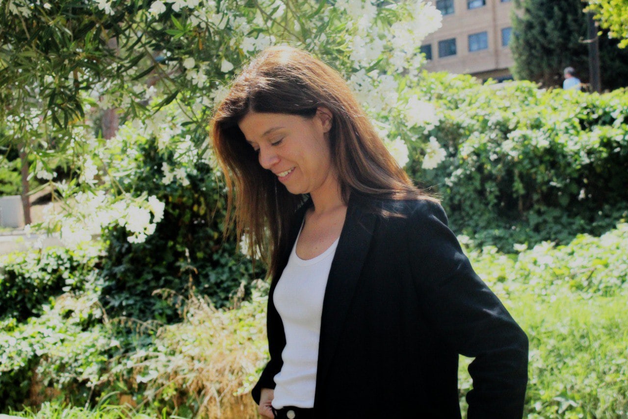 Side-profile of a smiling woman wearing a black blazer and white top, walking in a sunny garden with white flowers and greenery behind her.