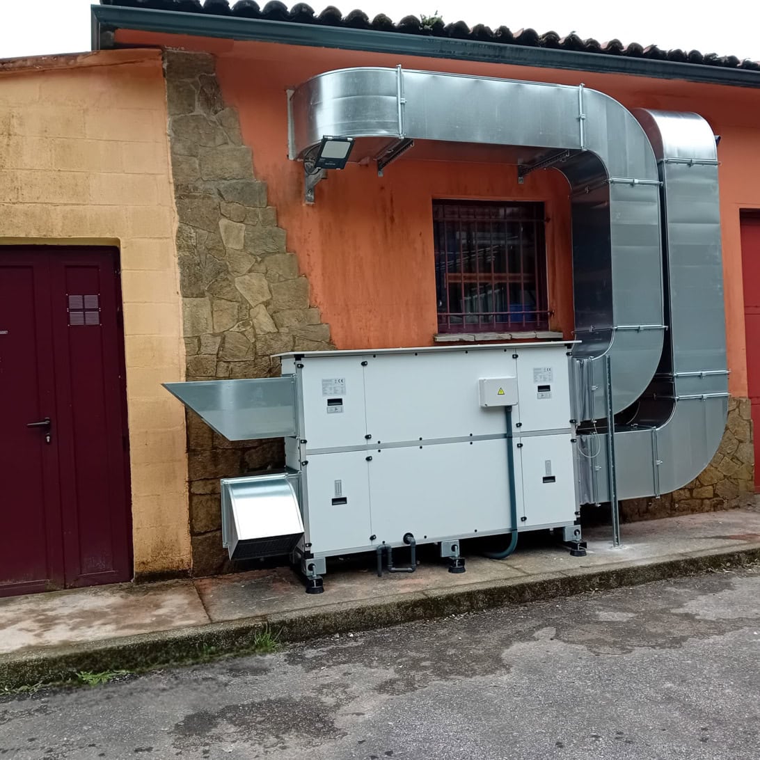 Outdoor industrial air handling unit with extensive galvanized ductwork mounted along an orange building wall, beside a barred window and a stone column on a paved surface.