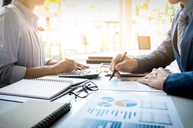 Two professionals reviewing charts and data on a desk, one using a calculator and the other writing with a pen.