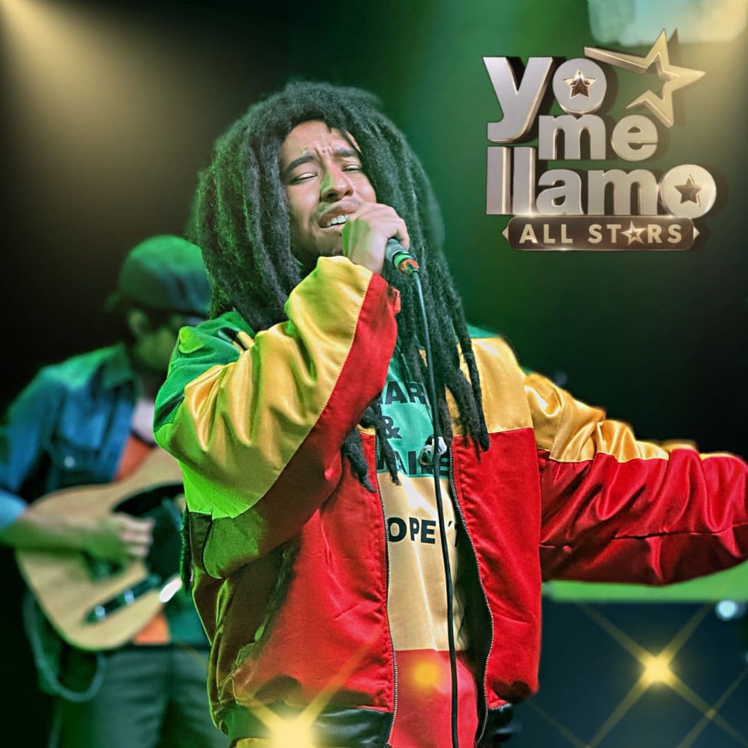 Male singer with dreadlocks performs on stage, wearing a bright red-yellow-green jacket and holding a microphone.