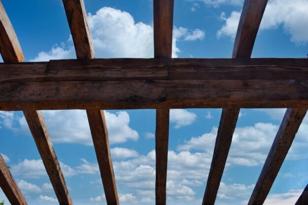 Wooden beam lattice framing a bright blue sky with white clouds, seen from below.