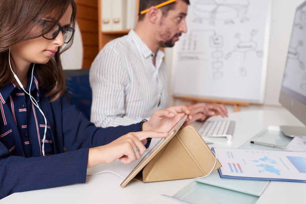 Two coworkers sit at a bright desk: a woman with glasses uses a tablet on a stand while a man types on a keyboard in the background, with a whiteboard of diagrams behind them.