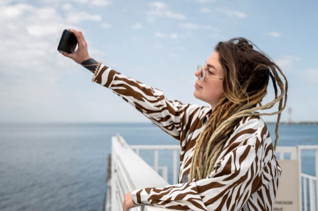 Woman with dreadlocks in a zebra-print shirt taking a selfie on a pier by the sea, wearing sunglasses.
