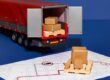 Model delivery truck with open rear doors showing stacked brown cardboard boxes on pallets inside the trailer, on a blue background next to a shipping map.