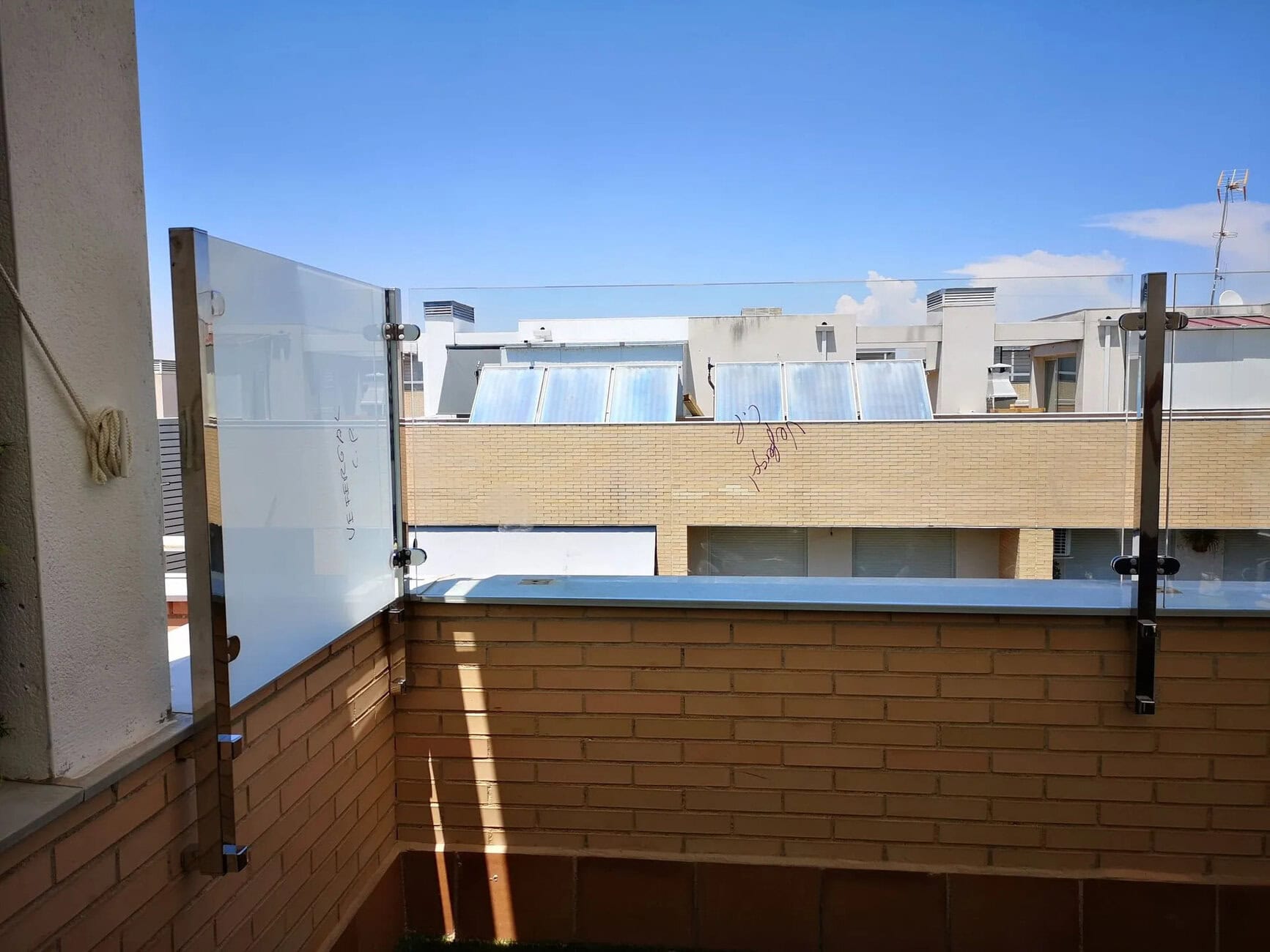 Balcony with glass railing off a brick wall, across a sunny blue sky and neighboring building roof with solar panels.