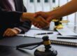 Handshake over a desk in a legal office, with a gavel, notebook, and laptop visible.