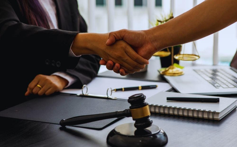 Handshake over a desk in a legal office, with a gavel, notebook, and laptop visible.
