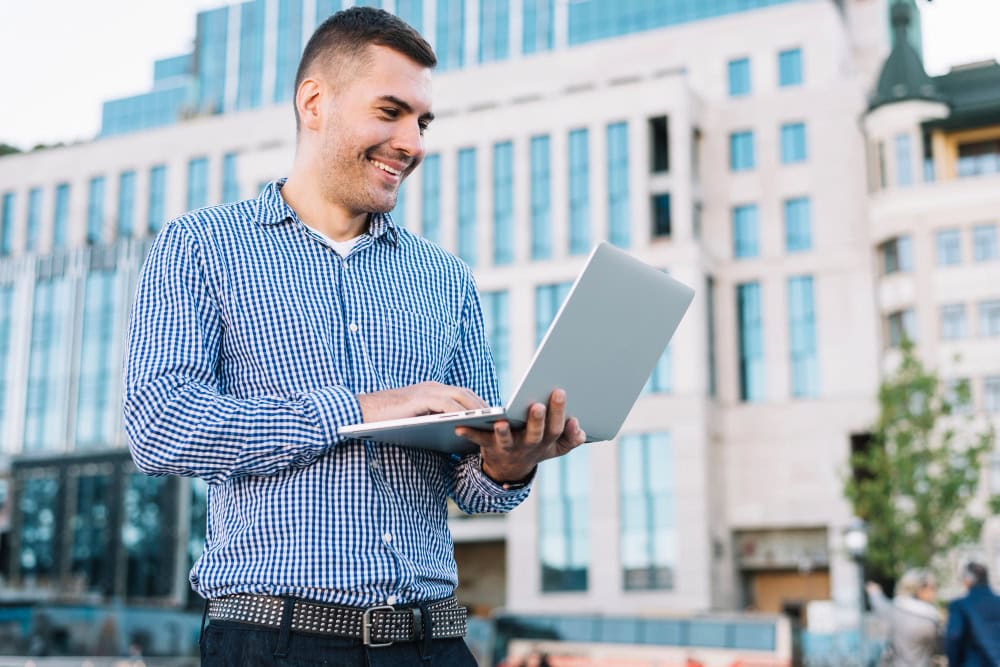 Man in a blue checkered shirt holding a silver laptop outdoors in front of a modern office building, smiling while using it.