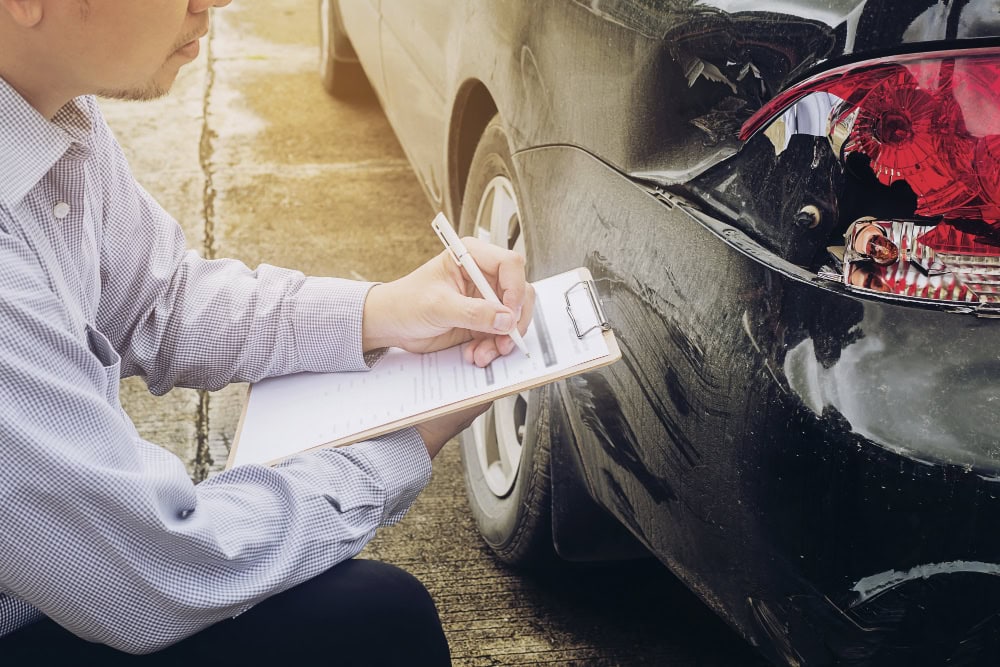 Man kneeling by a black car, writing on a clipboard to document damage to the rear bumper.
