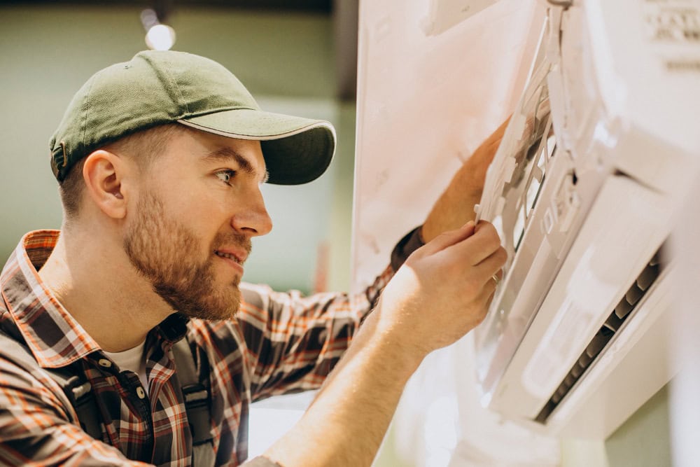 Man wearing a green cap and plaid shirt adjusting a wall-mounted machine's control panel with his right hand, likely performing maintenance.