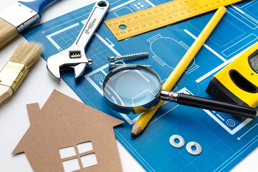 Architectural blueprint spread with tools: magnifying glass, adjustable wrench, pencil, yellow measuring tape, ruler, paintbrush, and a cardboard house cutout.