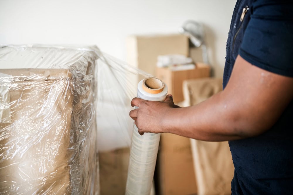 Person in a dark blue shirt wraps a large cardboard box with clear plastic stretch film in a warehouse setting.