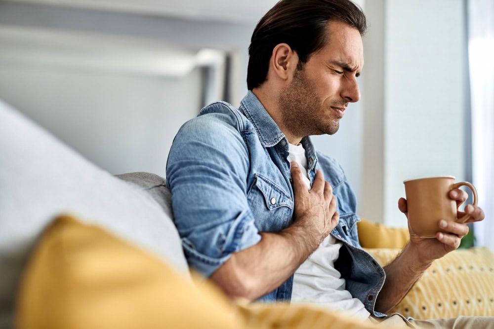 Man on a sofa clutches his chest in discomfort while holding a mug in his other hand.