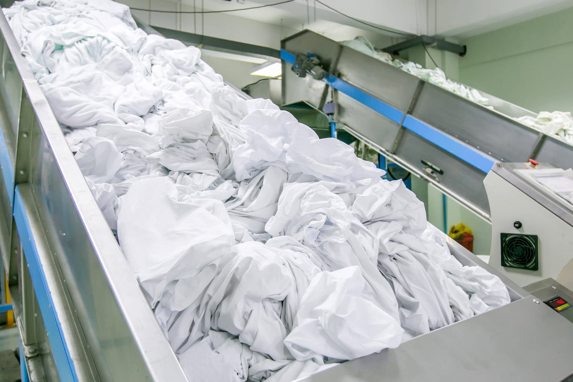 Industrial laundry conveyor transporting large piles of wrinkled white linens in a processing plant