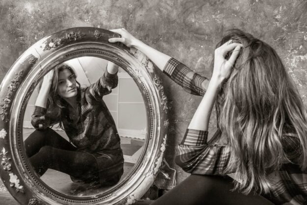 Woman in a plaid shirt sits on the floor, gazing at her reflection in a large ornate circular mirror.