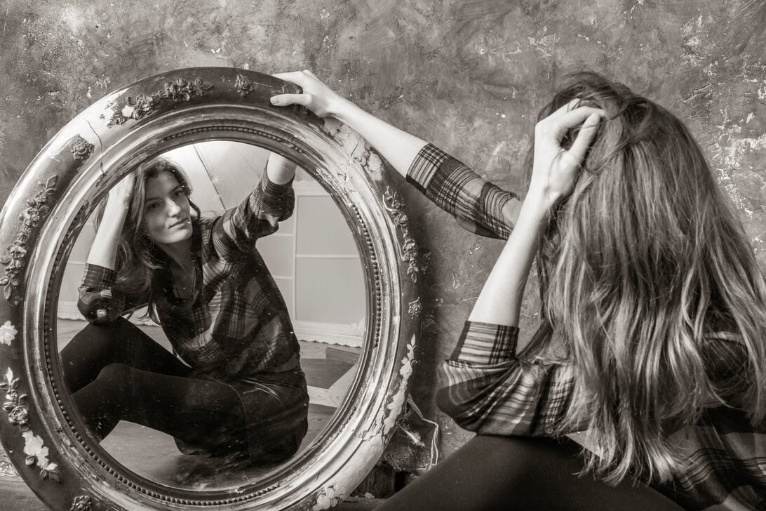 Woman in a plaid shirt sits on the floor, gazing at her reflection in a large ornate circular mirror.