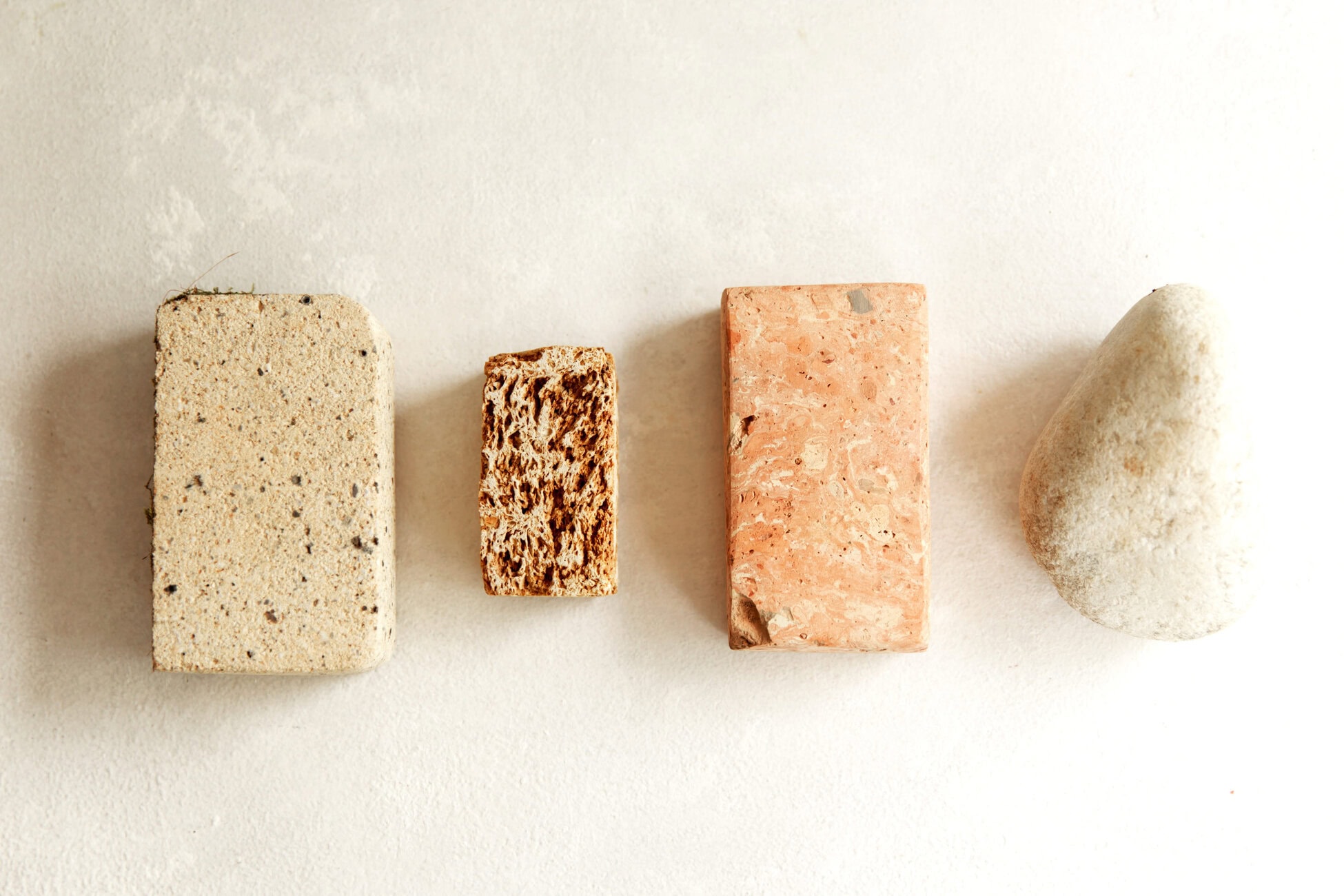 Four blocks lined up on a pale surface: a light speckled rectangular block, a small brown textured block, a pink speckled rectangular brick, and a smooth oval stone left to right.
