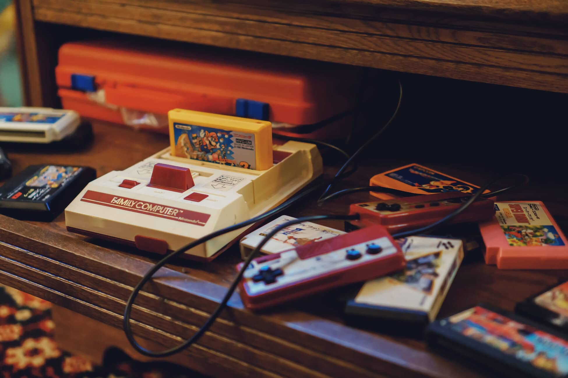 Retro Nintendo Famicom console with red controllers and colorful game cartridges on a wooden shelf, cables scattered nearby.