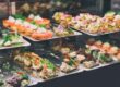 Assorted savory canapés and crostini on metal trays inside a bakery display case, garnished with herbs and vegetables.