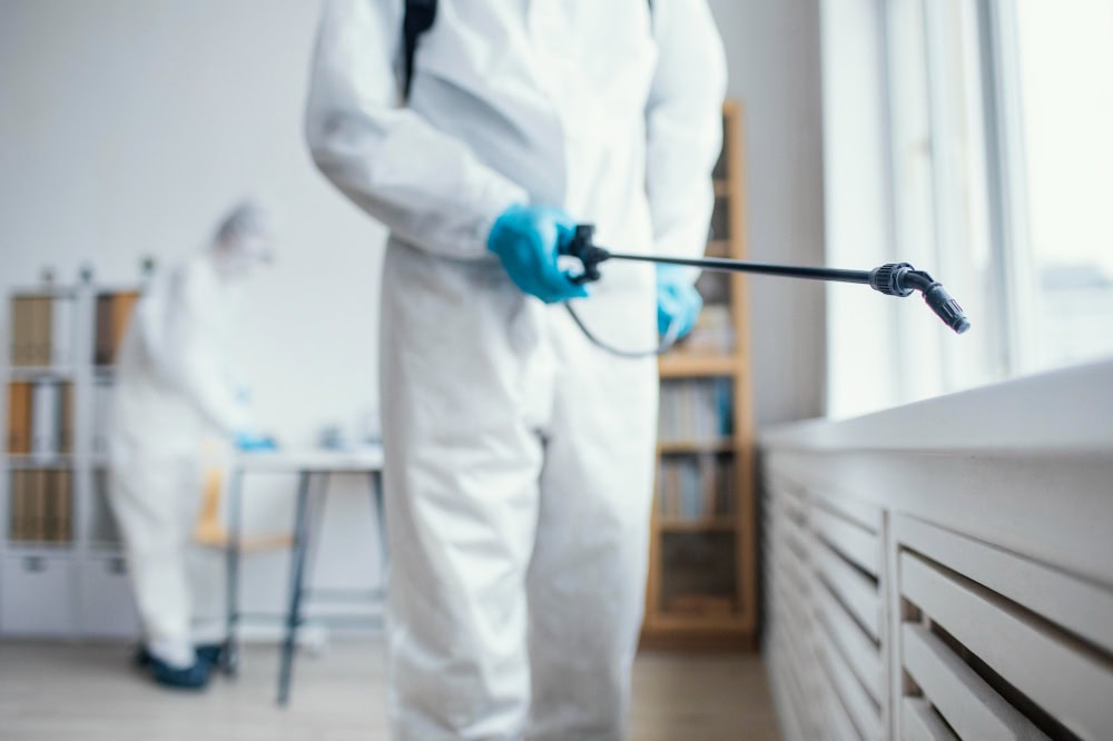 Worker in full protective suit spraying disinfectant with a long wand in a bright lab or clinic setting.