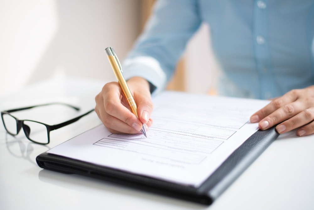 Person in a blue shirt writes on a form with a gold pen, with eyeglasses resting on the desk nearby