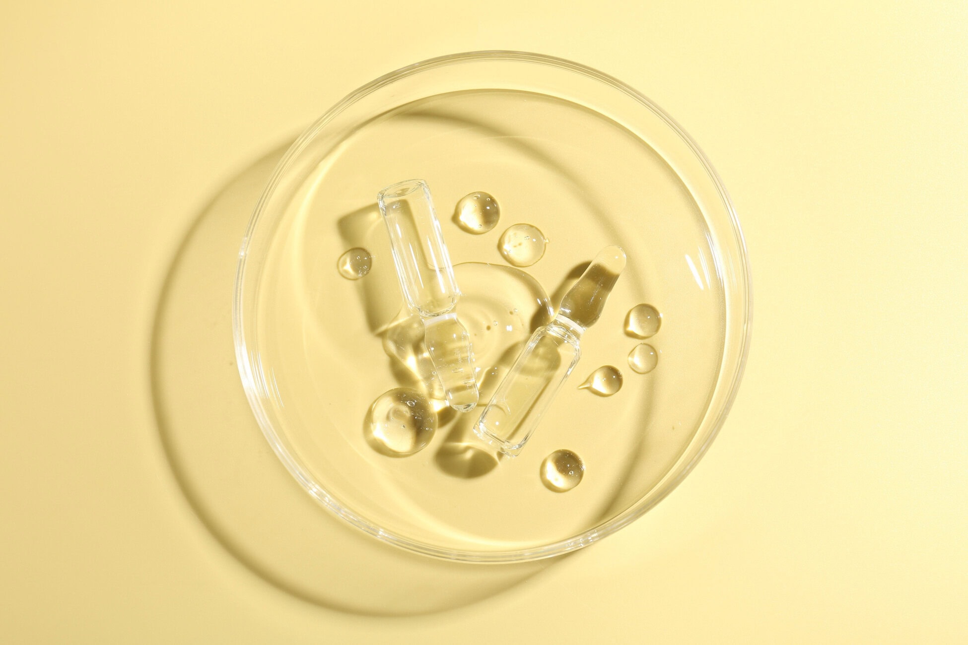 Top-down close-up of a glass bowl with ice cubes and droplets on a pale yellow surface.