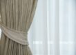 Beige drapes pulled to the side, revealing sheer white curtains over a window, letting light in.