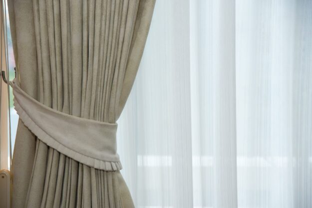 Beige drapes pulled to the side, revealing sheer white curtains over a window, letting light in.