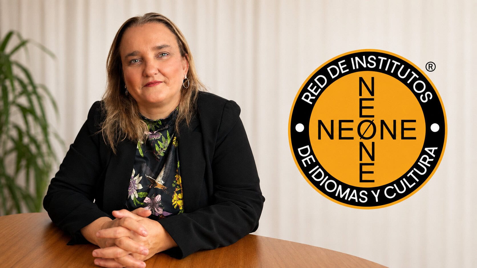 Mujer sentada junto a una mesa de madera, wearing blazer negro, frente a un logotipo circular naranja y negro de la Red de Institutos de Idiomas y Cultura.