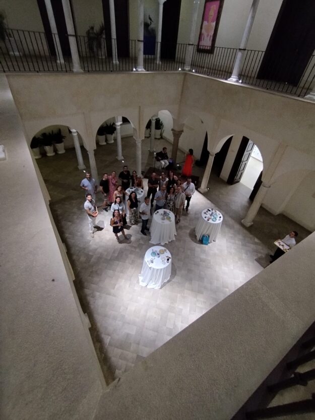 A group of about 20 people standing in a circle around tall round tables with white tablecloths in a stone atrium, viewed from a balcony; several people wear light clothing and a woman in a red dress stands near the center.