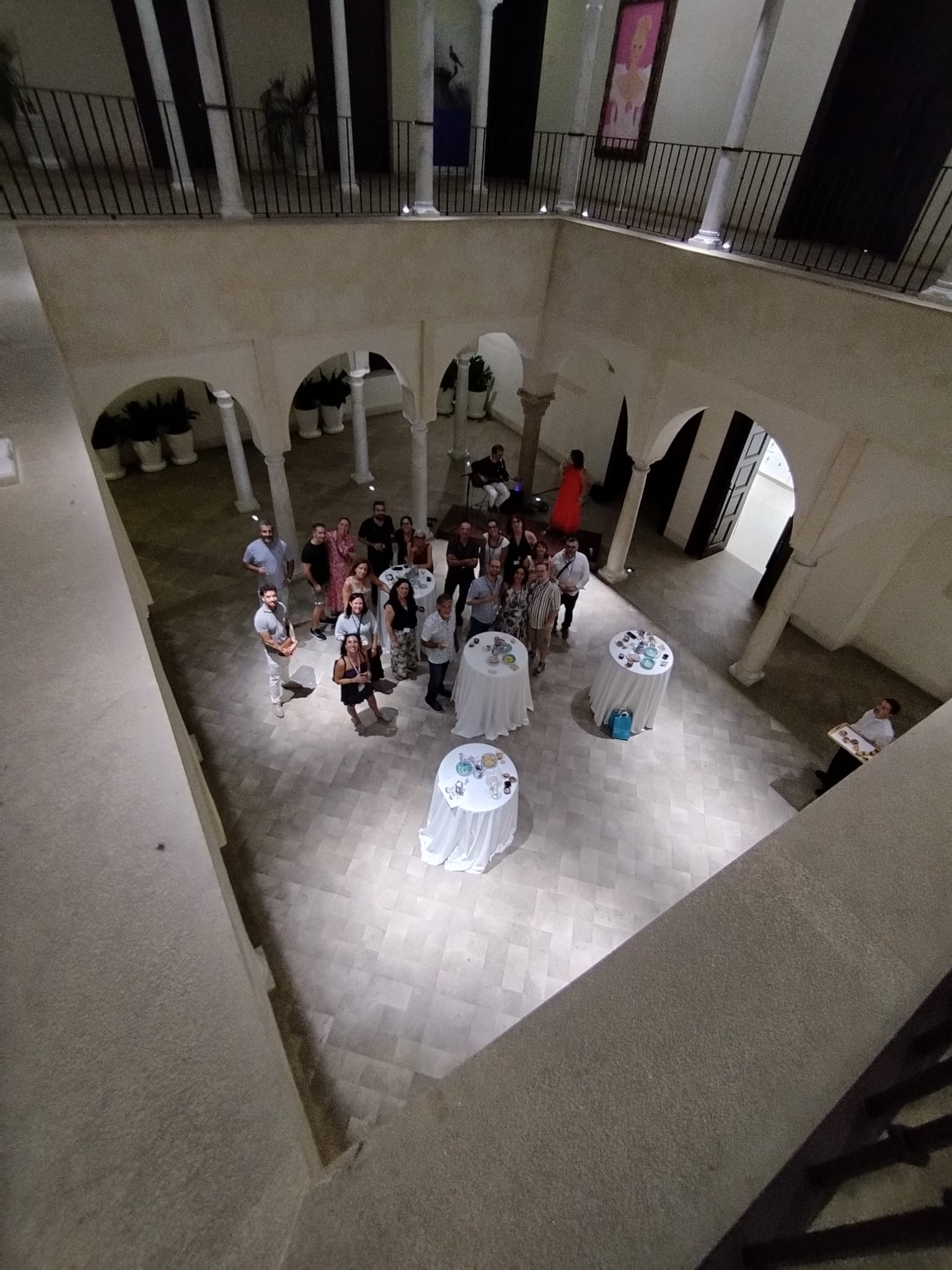 A group of about 20 people standing in a circle around tall round tables with white tablecloths in a stone atrium, viewed from a balcony; several people wear light clothing and a woman in a red dress stands near the center.